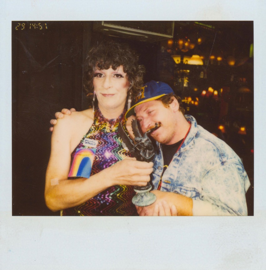 Download the full-sized image of A Photograph of Sylvia Rivera Wearing a Colorful Sequined Dress, Holding a Statuette, and Being Hugged By Another Person