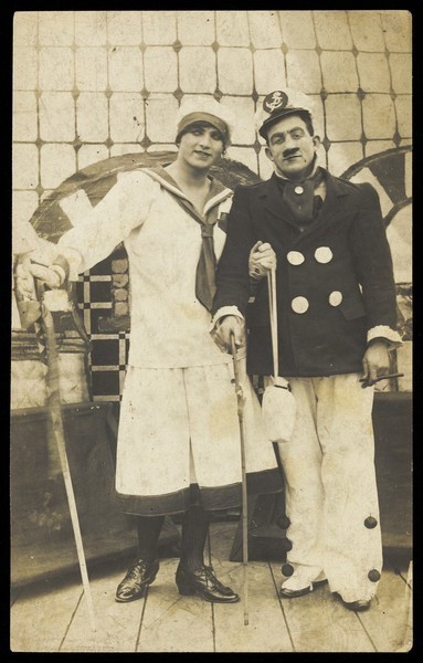Download the full-sized image of Two sailors, one in drag, pose on deck holding props. Photographic postcard, 191-.
