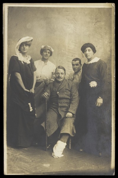Download the full-sized image of Four soldiers posing, three in drag, with an injured soldier seated in the centre. Photographic postcard, 191-.