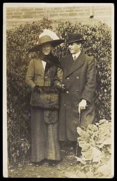 Download the full-sized image of Two men, one in drag, wearing smart attire, pose as a couple within foliage. Photographic postcard, ca. 1900.