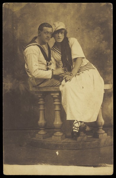 Download the full-sized image of Two sailors, one in drag, embracing while sitting on a stage balcony. Photographic postcard, 191-.