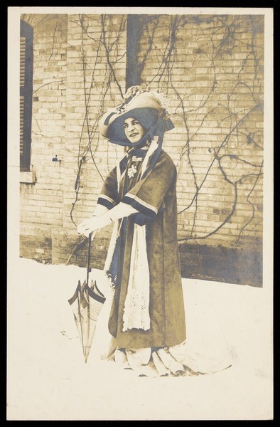 Download the full-sized image of A man in drag, wearing a large bonnet, leans on a parasol whilst standing in front of a brick building. Photographic postcard, 191-.