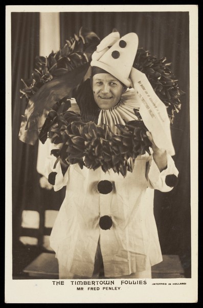 Download the full-sized image of Fred Penley dressed as a clown holding a wreath, performing for "The Timbertown Follies" at a prisoner of war camp in Groningen. Photographic postcard, 191-.