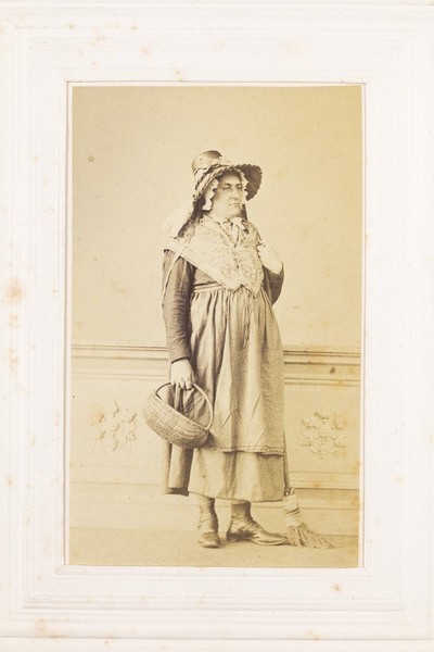 Download the full-sized image of A man in drag poses with a bonnet, basket and broom. Photograph, 189-.