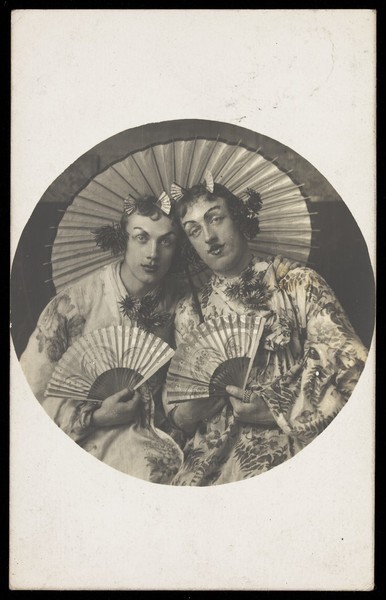 Download the full-sized image of Two young men in drag wearing Japanese costumes, pose together holding fans, underneath a parasol. Photographic postcard, 1905.