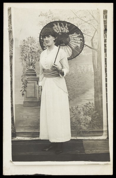 Download the full-sized image of An amateur actor in drag poses with an open parasol, in front of painted scenery. Photographic postcard, 191-.