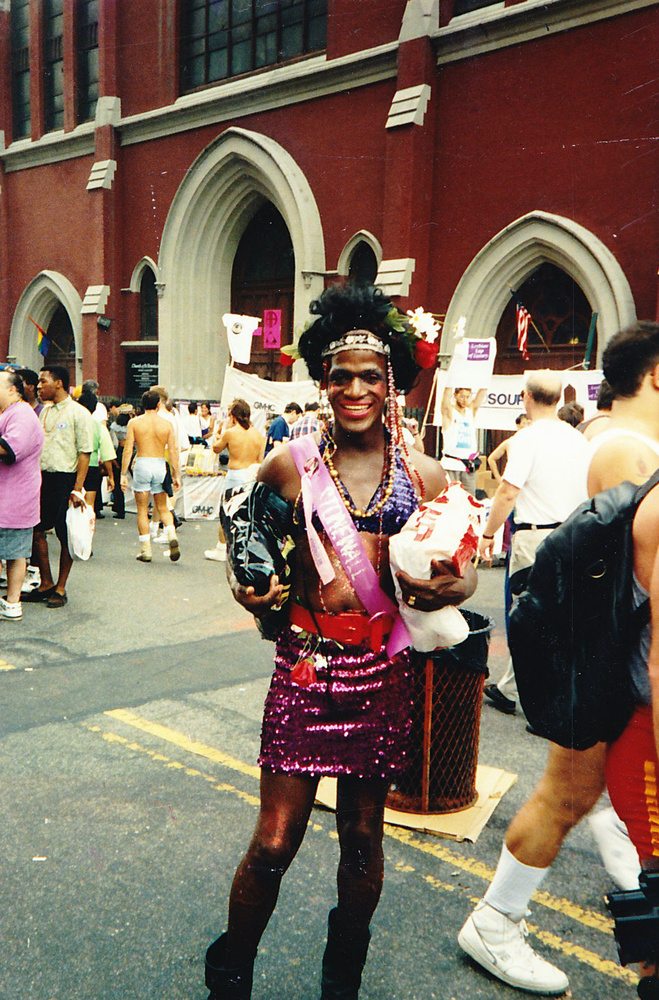 Download the full-sized image of A Photograph of Marsha P. Johnson Holding Bags, Wearing a Sequined Skirt and a Pink “Stonewall” Sash